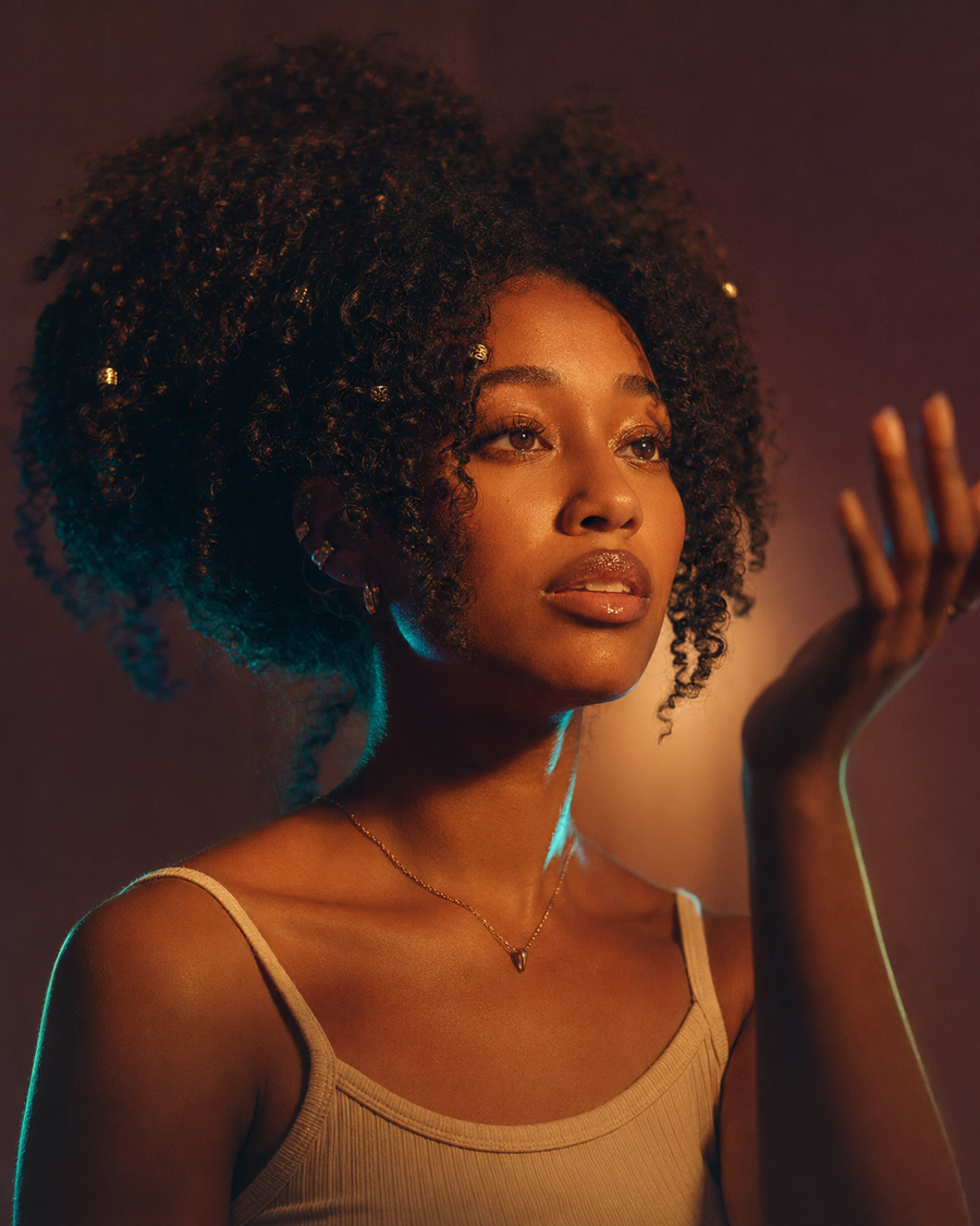 Amara portrait — warm amber light on the right, cool teal rim on the left, close-up: natural curls with tiny gold beads, hand raised into a soft halo of light, thin gold pendant necklace. Grounded, radiant, searching.