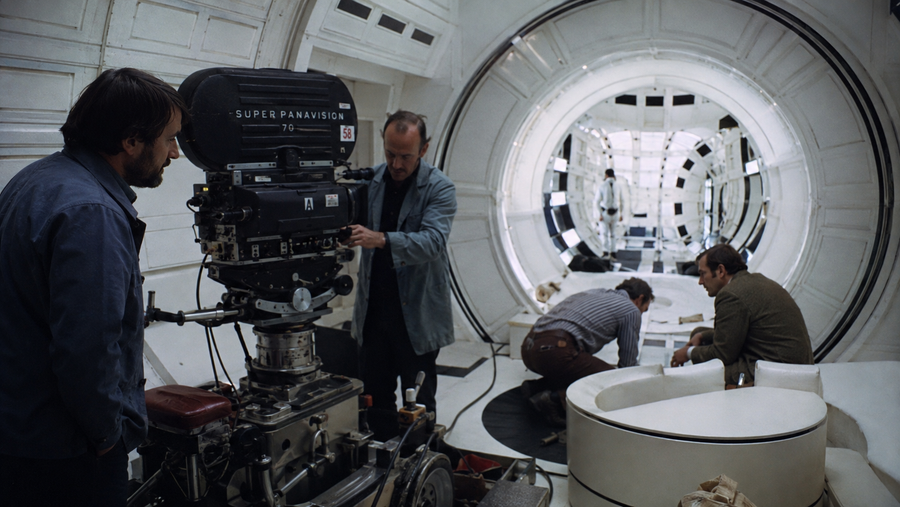GREYLIGHT — behind the scenes on a 1968-style sci-fi set: Super Panavision 70 camera aimed down a white space-station corridor. The machinery the world of In the Blink of 2073 is built from.