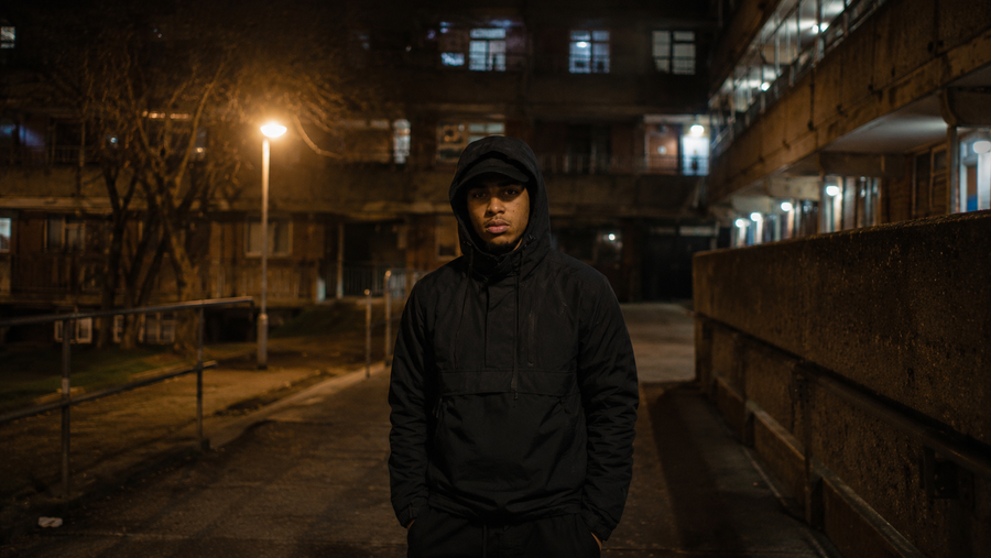 Static Twin — alone on a sodium-lit council-estate walkway at night, tower blocks behind him. The world of Who's Real (Duality Version).